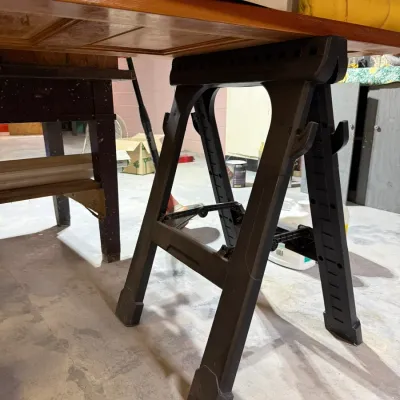 Black plastic folding sawhorse supporting a wooden tabletop in a basement workshop.