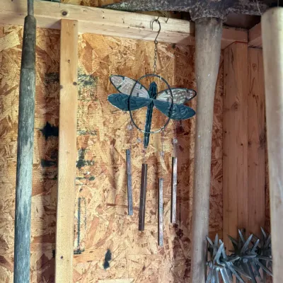 Decorative metal dragonfly wind chime hanging in a shed alongside vintage garden tools, including a pickaxe and a hoe.