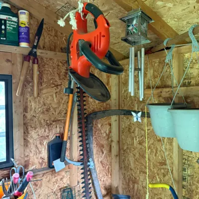 Black & Decker electric hedge trimmer hanging in a garden shed alongside various gardening tools, wind chimes, and hanging planters.