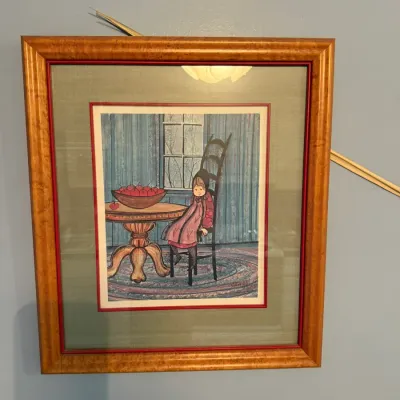 Framed folk art print depicting a young girl seated at a table with a bowl of apples, presented in a warm wood frame with double matting.