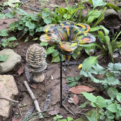 Hand-blown glass flower garden stake with vibrant yellow, orange, and green swirls, displayed alongside a small concrete pinecone finial.