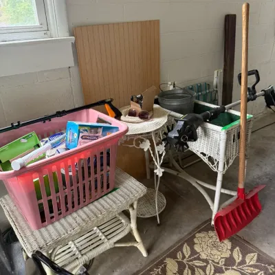 Assorted household and garden items, including white wicker patio furniture, a pink laundry basket filled with cleaning supplies, a red...
