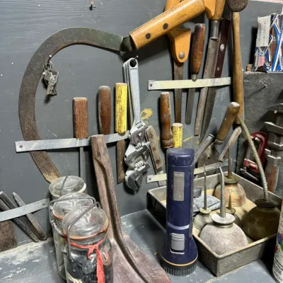 Assorted vintage hand tools, including a sickle, hand saws, files, adjustable wrenches, and oil cans, arranged on a workbench and pegboard.