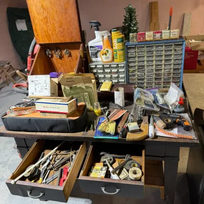 Vintage wooden workbench with open drawers, featuring an assortment of hand tools, hardware organizers, cleaning supplies, and a wooden...