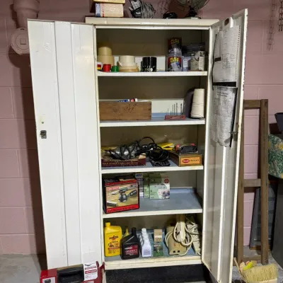 Vintage white metal utility cabinet with double doors, filled with various workshop tools, hardware, and household supplies.