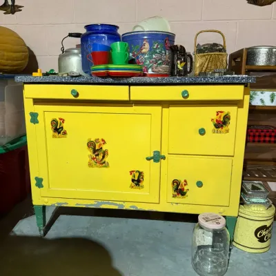 Vintage yellow-painted Sellers-style kitchen cabinet featuring green hardware and rooster decals, topped with various kitchenware items.