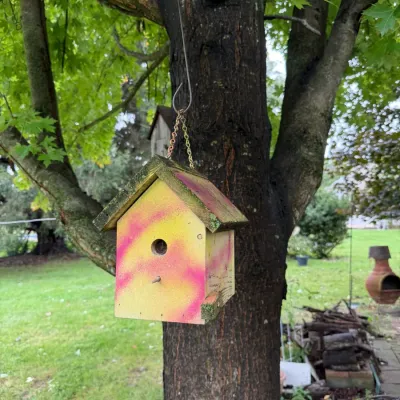Handmade wooden birdhouse painted with yellow and pink spray paint, hanging from a tree branch by a metal chain.