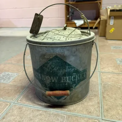 Vintage J.C. Higgins galvanized metal minnow bucket with a green logo, wire handle, and wooden grip, showing age-appropriate wear and...