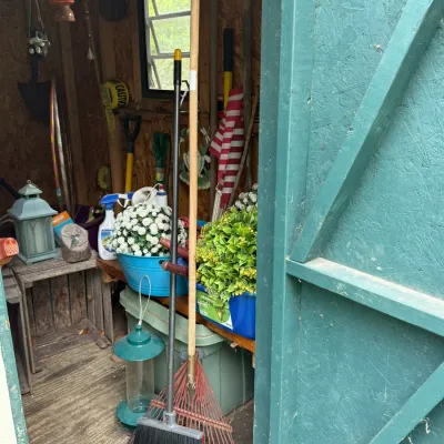 Assorted garden tools, planters, and outdoor decor stored inside a wooden shed, including a broom, leaf rake, bird feeder, and artificial...
