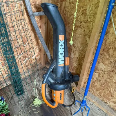 Worx electric leaf blower and mulcher, shown stored in a shed alongside garden tools and wire fencing.