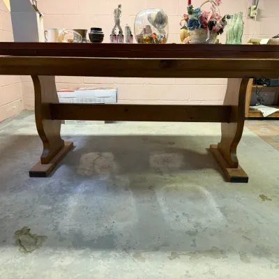 Rustic pine trestle-style dining table with curved support legs and a central stretcher, displaying various small collectibles on top.