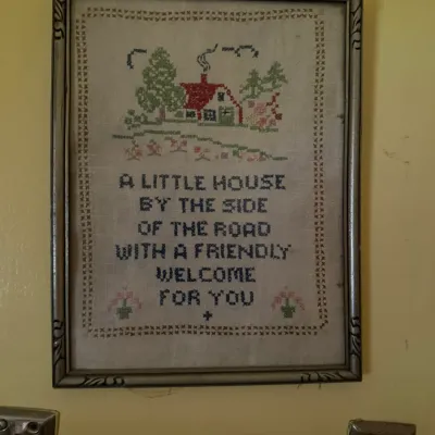 Vintage framed cross-stitch sampler featuring a house illustration and the quote "A little house by the side of the road with a...