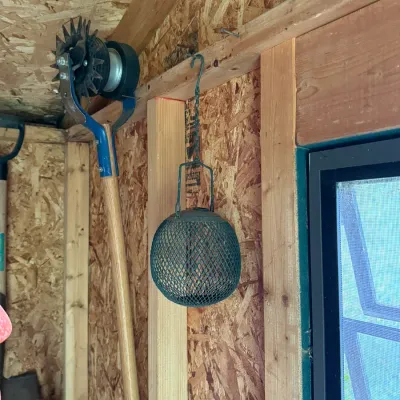 Vintage green wire mesh spherical bird feeder hanging from a hook, alongside a rotary lawn edger with a wooden handle.