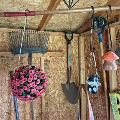 Assorted garden tools and outdoor decor hanging in a shed, including a metal rake, a shovel, a hanging artificial flower ball, and a...