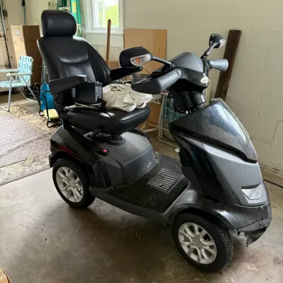 Four-wheel mobility scooter in black and grey, featuring a high-back captain's chair, side mirrors, and a battery charger resting on...