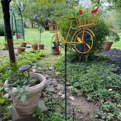 Whimsical yellow metal bicycle wind spinner garden stake with colorful pinwheel spokes and front and rear baskets.
