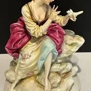 Capodimonte-style porcelain figurine depicting a seated woman with a bird perched on her hand, featuring detailed hand-painted clothing...