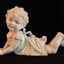 Vintage bisque porcelain piano baby figurine depicting a crawling child in a nightshirt with a blue bow, in good condition.
