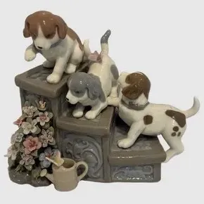 Vintage porcelain figurine depicting three playful puppies on garden steps, featuring delicate floral details and a small watering can....