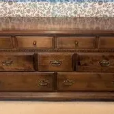 Vintage dark wood triple dresser featuring nine drawers with brass bail pulls and small knob hardware, set against a floral wallpaper...