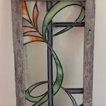 Stained glass window panel featuring a stylized floral design with green and orange accents, set in a rustic weathered wood frame with...