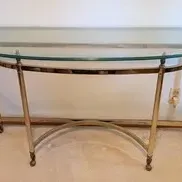Hollywood Regency style demilune console table featuring a brass-finished metal frame with curved stretchers and a thick glass top.