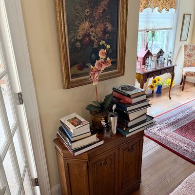 Vintage wooden cabinet with a framed floral oil painting, stacks of books, and a faux orchid arrangement.