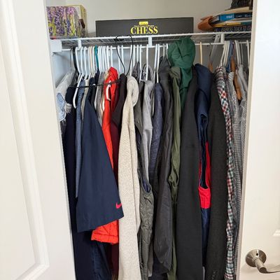 Assorted men's outerwear, shirts, and jackets hanging in a closet, with a boxed chess set and other items stored on the wire shelf...
