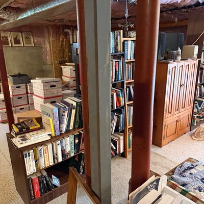 Unfinished basement filled with various items for sale, including multiple bookshelves packed with books, cardboard storage boxes, and a...