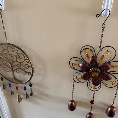 Two decorative metal wind chimes hanging on a wall, one featuring a tree of life design with small colorful beads and bells, and the...