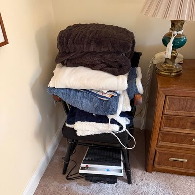 Stack of assorted blankets and throws in brown, white, and blue, piled on a black wooden chair next to a Medi-Rub foot massager.