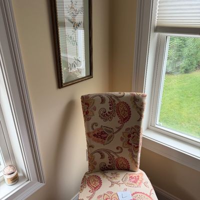 Upholstered dining or accent chair featuring a warm-toned paisley floral pattern, shown alongside a framed botanical print on the wall.