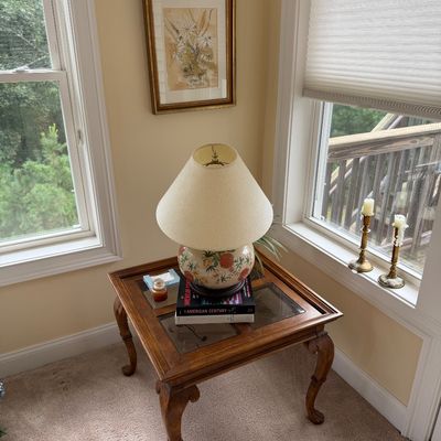 Traditional wood end table with cabriole legs and glass-insert top, featuring a floral ceramic table lamp and framed botanical artwork.