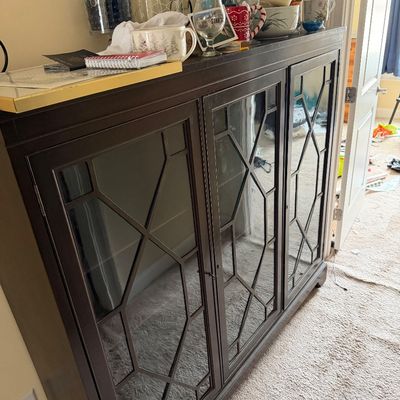 Contemporary dark wood sideboard featuring three glass-front doors with geometric mullion overlays, topped with various glassware and...