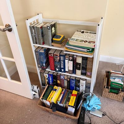 Small white wooden three-shelf bookcase filled with various history and reference books, accompanied by additional books in woven baskets...
