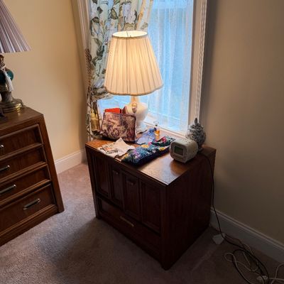 Mid-century modern style wooden nightstand featuring a white ceramic table lamp with a pleated shade, alongside small personal...