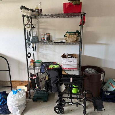 Four-tier wire shelving unit on casters, holding various household items, alongside a black folding transport wheelchair.