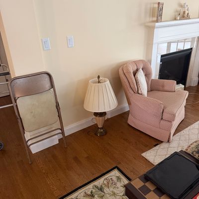 Vintage pink upholstered armchair with a tufted back, paired with a traditional table lamp featuring a pleated shade, and a metal folding...