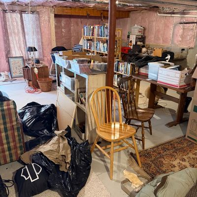 Unfinished basement filled with various estate sale items, including wooden Windsor-style chairs, bookshelves, storage bins, and...
