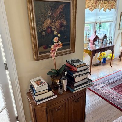 Traditional wooden two-door cabinet displaying stacks of books, an artificial orchid, and a framed floral oil painting hanging above.