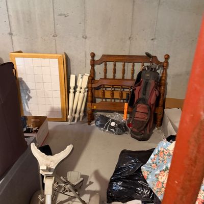 Assorted household items in a basement, including a wooden spindle headboard, a golf bag with clubs, a pull-behind golf cart, and...