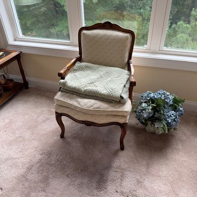 Vintage French Provincial style armchair with carved wood frame and cream upholstery, shown with a folded green quilt and a faux blue...