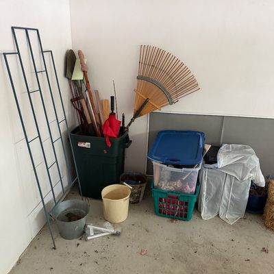 Assorted garden and garage items, including a blue metal trellis, a bamboo rake, a green trash bin holding various tools, plastic storage...