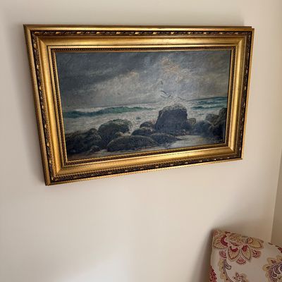Vintage oil painting depicting a stormy seascape with waves crashing against a rocky shoreline, housed in an ornate, gilded frame.