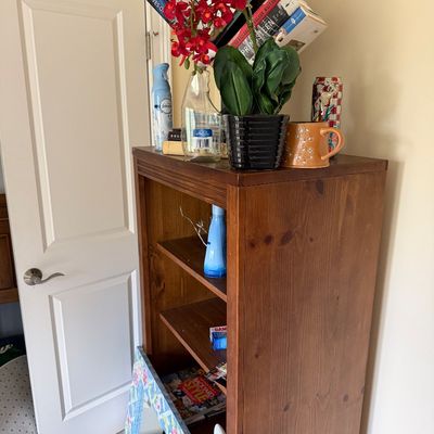 Mid-sized wooden bookcase featuring adjustable shelves, topped with books, artificial red orchids, and decorative mugs.