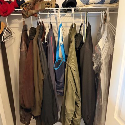 Assorted men's outerwear, including leather and utility jackets, hanging in a closet alongside a blue tote bag and folded blankets.