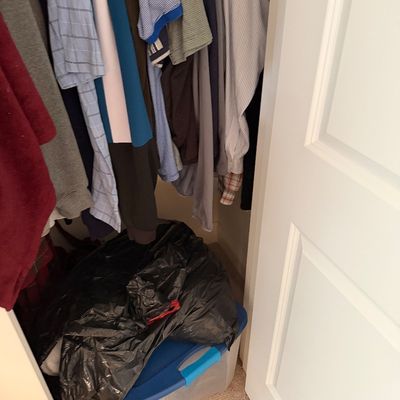 Assorted men's clothing hanging in a closet, including polo shirts and button-downs, above a plastic storage bin and a black trash bag.