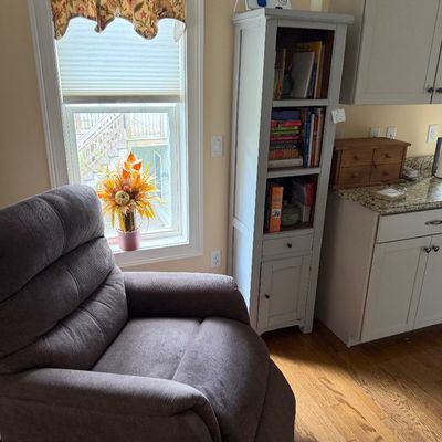 Plush grey fabric recliner armchair positioned next to a tall, narrow grey-painted wooden bookcase with a lower cabinet.