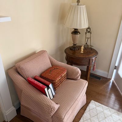 Upholstered armchair in a dusty rose patterned fabric, paired with a round wooden side table holding a traditional table lamp and...