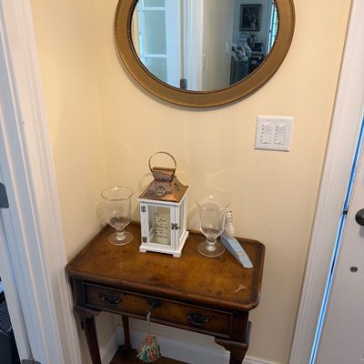 Vintage Queen Anne style wood side table with a single drawer, paired with a round gold-framed wall mirror and decorative tabletop...